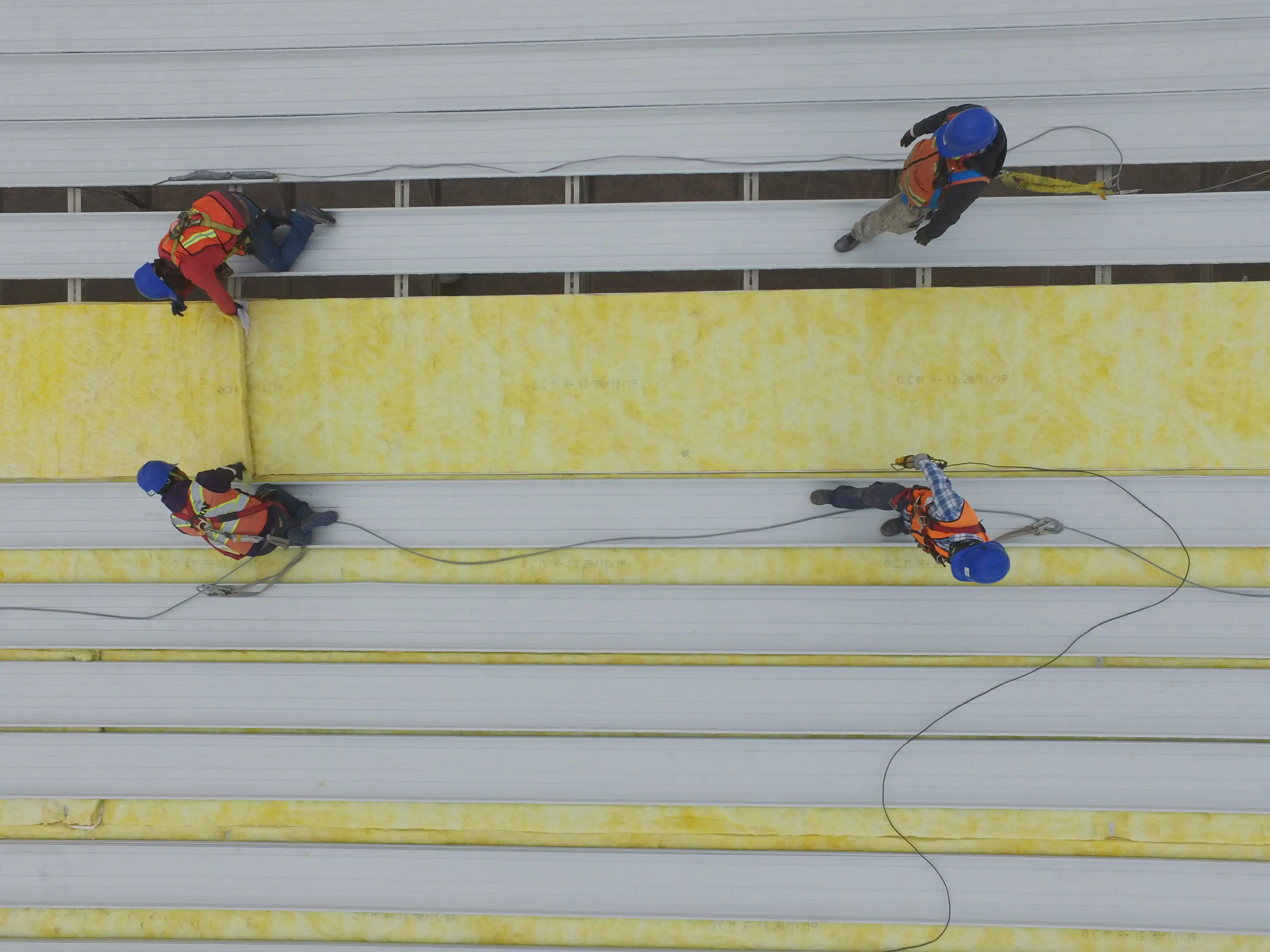 Vista aérea de cuatro trabajadores de la construcción con arneses y equipo de seguridad instalando aislamiento térmico en un techo, ejemplificando la prevención de riesgos laborales y la gestión de riesgos en construcción.