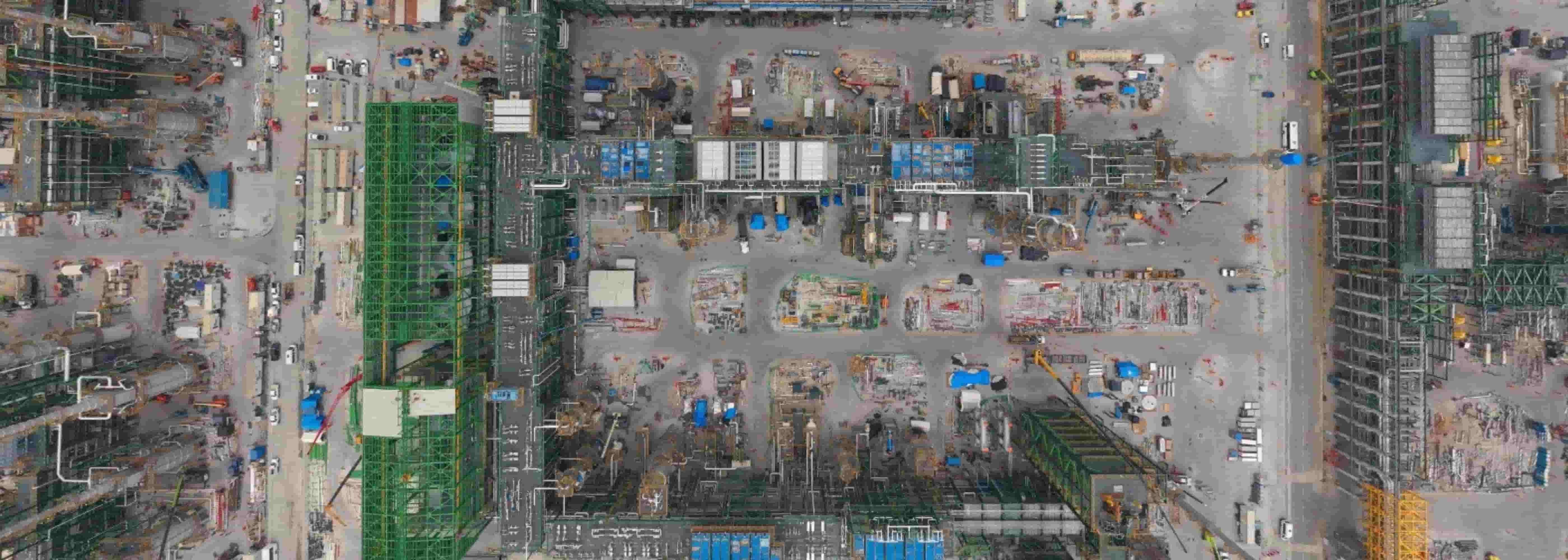 Vista aérea cenital de un vasto sitio de construcción industrial complejo o una planta de procesamiento masiva. Se observan estructuras de acero verdes, edificios grises, numerosas tuberías y maquinaria en el suelo, representando un proyecto de construcción a gran escala como un Data Center. 