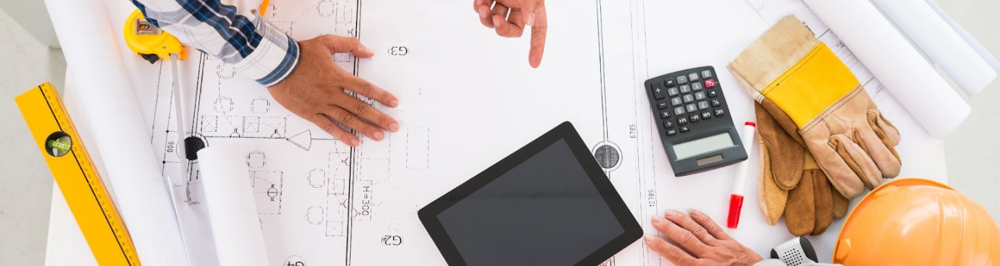 Vista superior de planos arquitectónicos con herramientas de construcción, tablet y equipo de protección, mientras dos personas discuten detalles técnicos, simbolizando la planificación colaborativa en proyectos EPC.