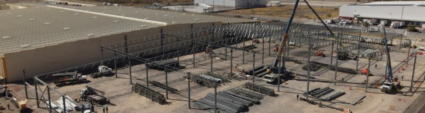 Gran nave industrial en proceso de construcción con estructura metálica de acero, grúas amarillas y personal trabajando en el sitio bajo un cielo despejado.