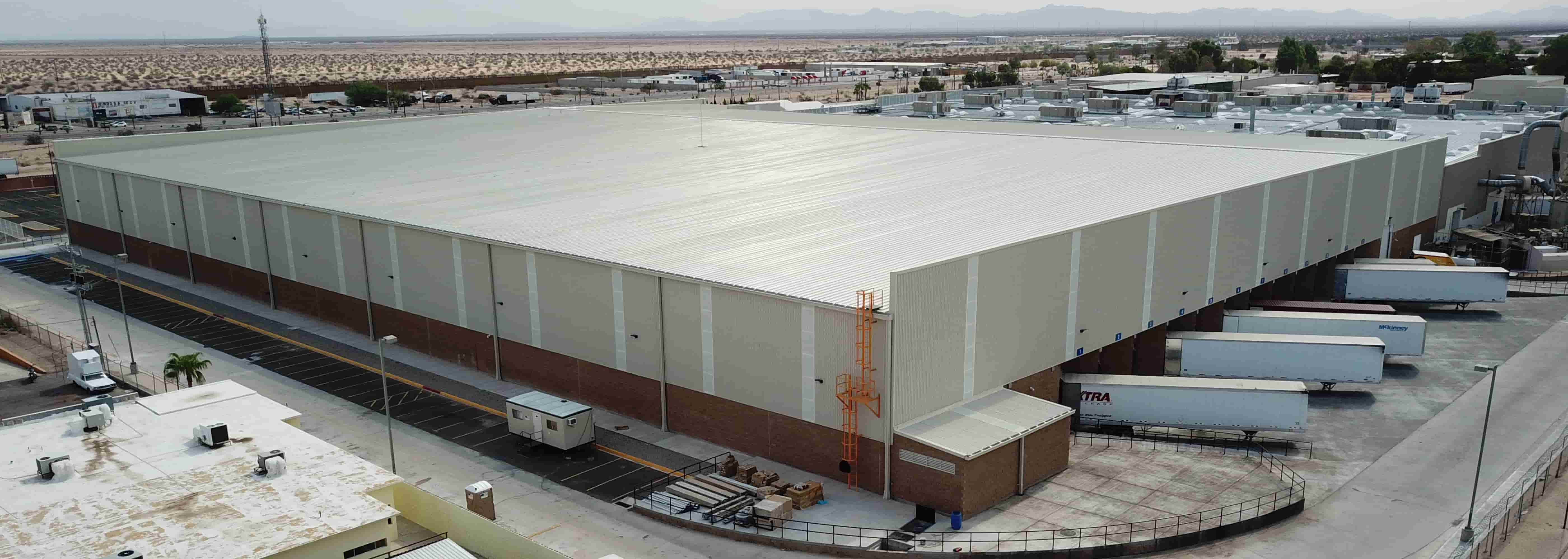 Vista aérea de un gran almacén o nave industrial moderna de estructura metálica, con techo de color claro y muros combinados de metal y ladrillo, ubicada en una zona semiárida. La instalación incluye una zona de carga y descarga con tráileres, ilustrando un proyecto de construcción industrial donde se aplica la Ingeniería de Valor para optimizar costos.